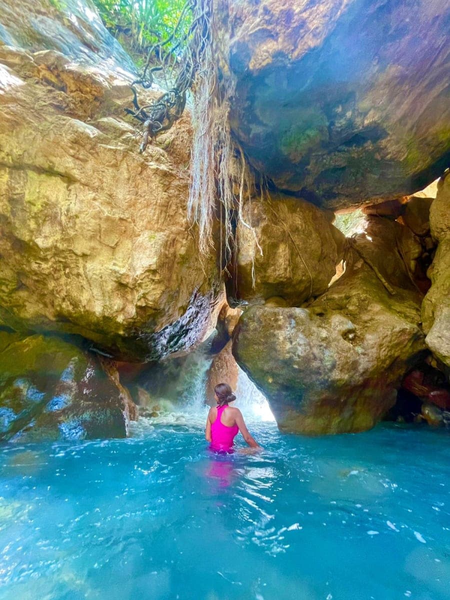 A swimmer in turquoise water inside a rock canyon below a thin waterfall